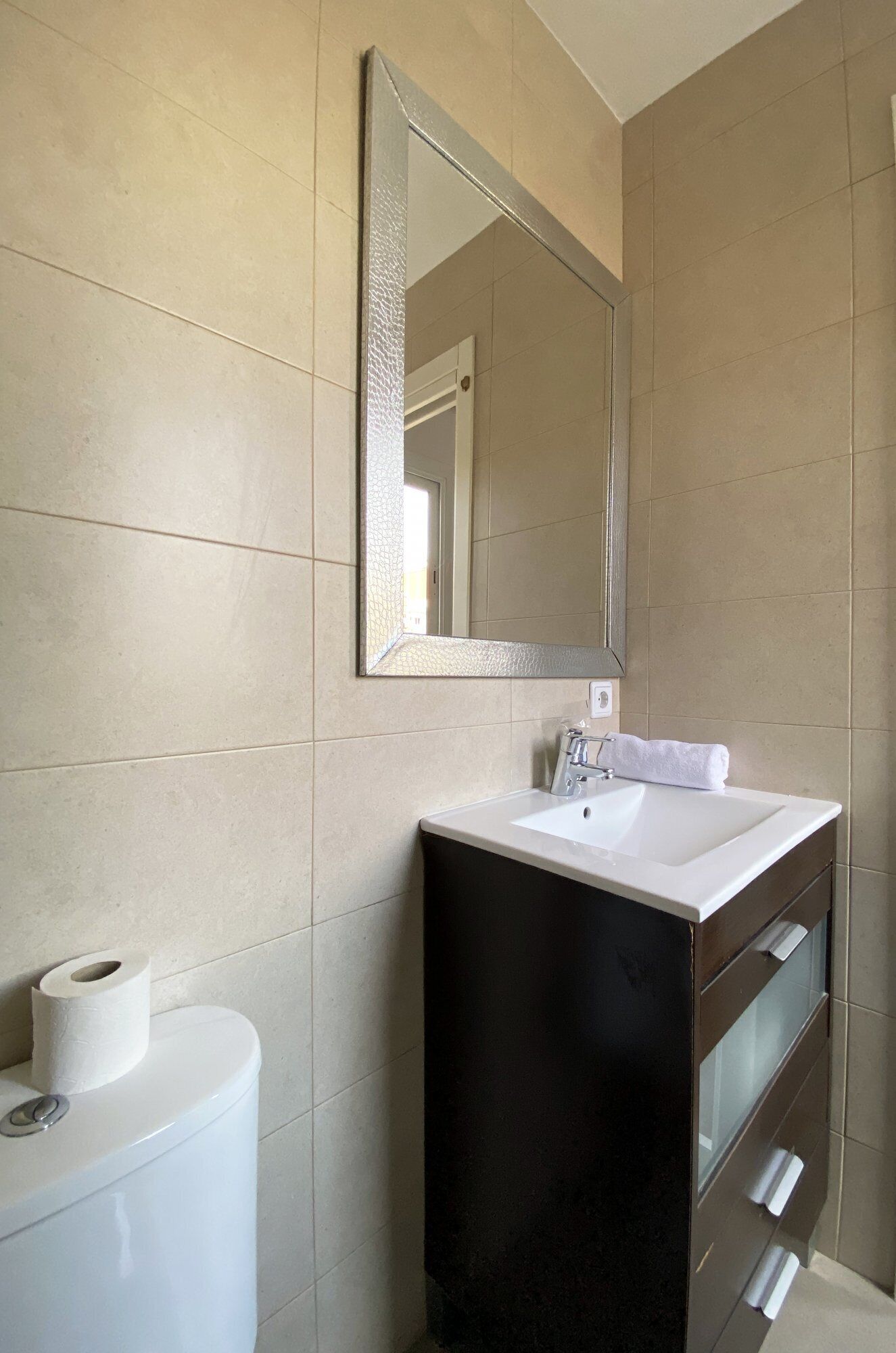 Une salle de bain moderne avec des toilettes blanches, un lavabo blanc et un meuble sombre en dessous, ainsi qu’un miroir au-dessus du lavabo.