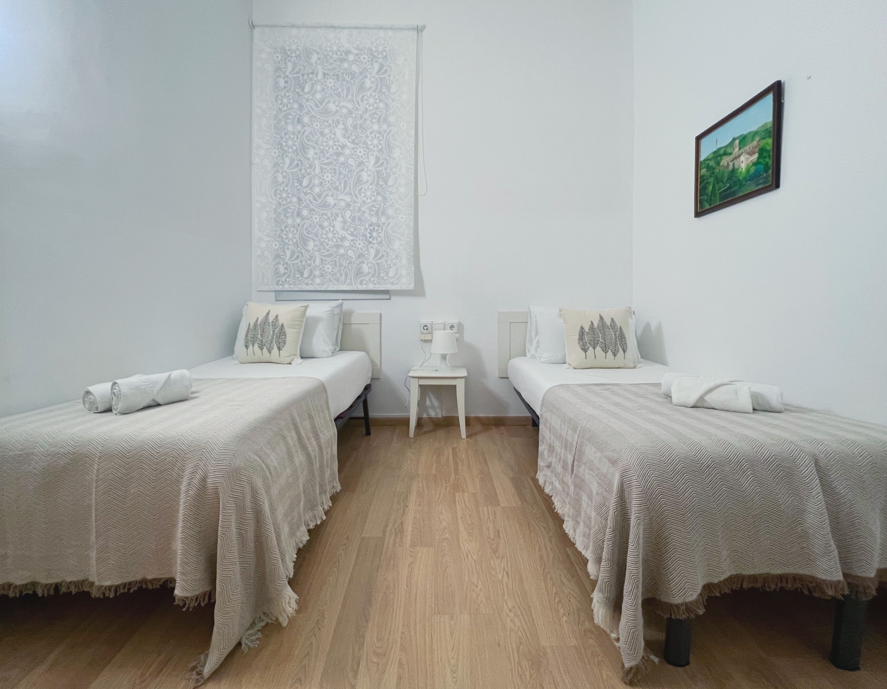 Une chambre avec deux lits, une petite table, une fenêtre avec un rideau léger et un tableau encadré au mur.