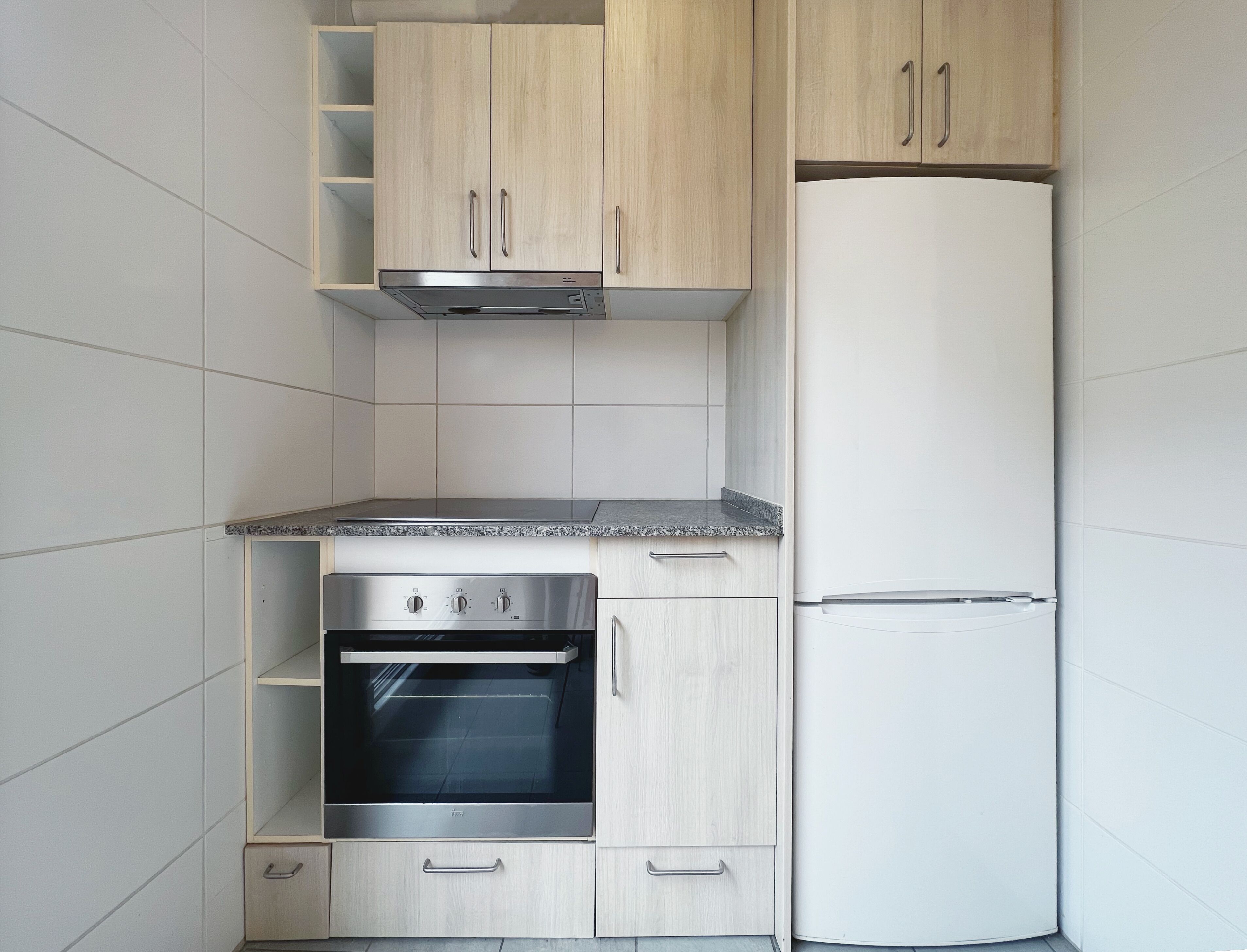 Une cuisine compacte dotée d’un four encastré, d’un réfrigérateur et de meubles de rangement en bois clair.