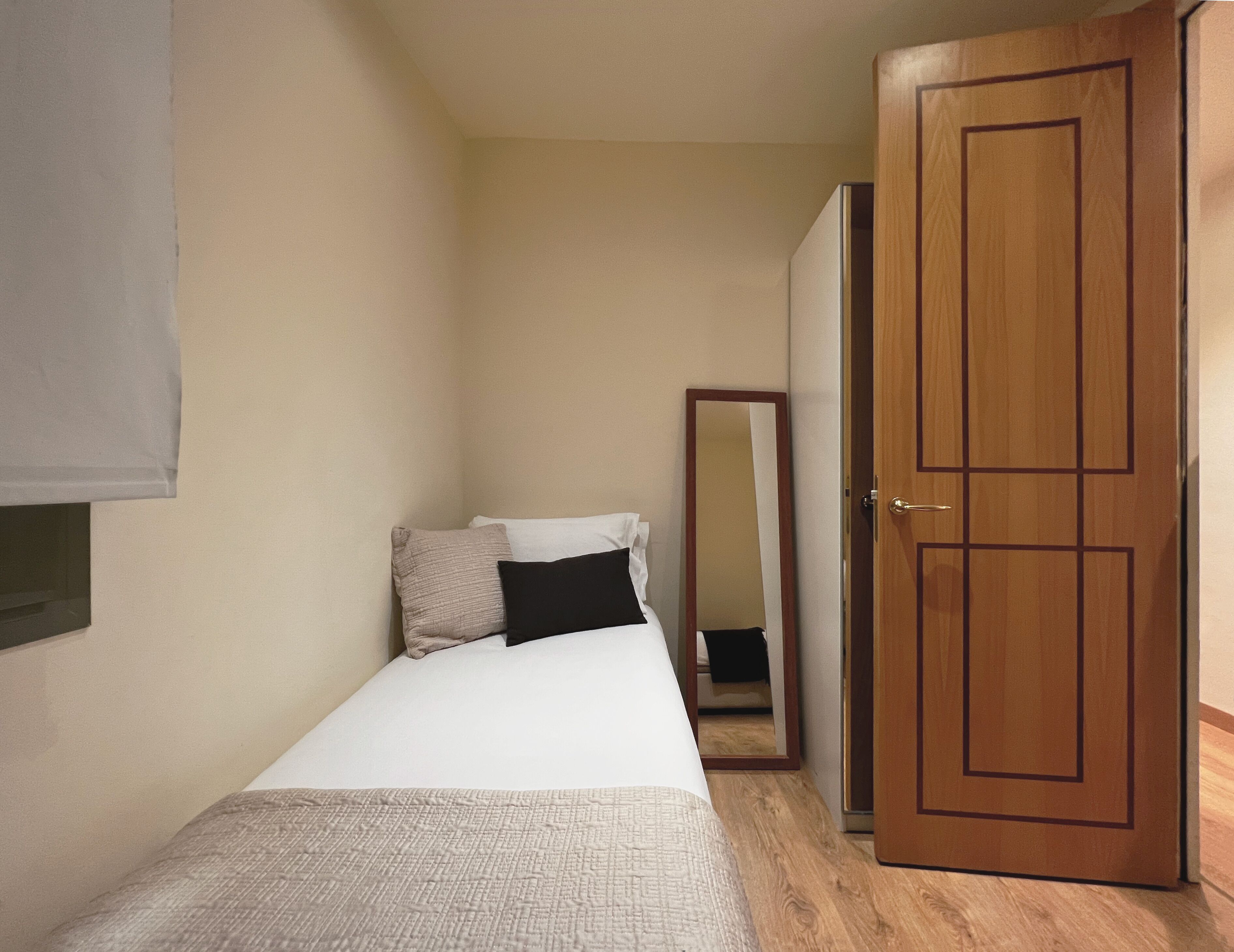 A small, neatly arranged bedroom with a single bed, a wooden wardrobe, and a mirror on the wall.
