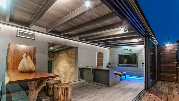 Ein moderner Außenbereich mit Küche, einem Holztisch, Hockern, einer Ziegelwand und einem großen Fenster mit Blick auf die Terrasse.
