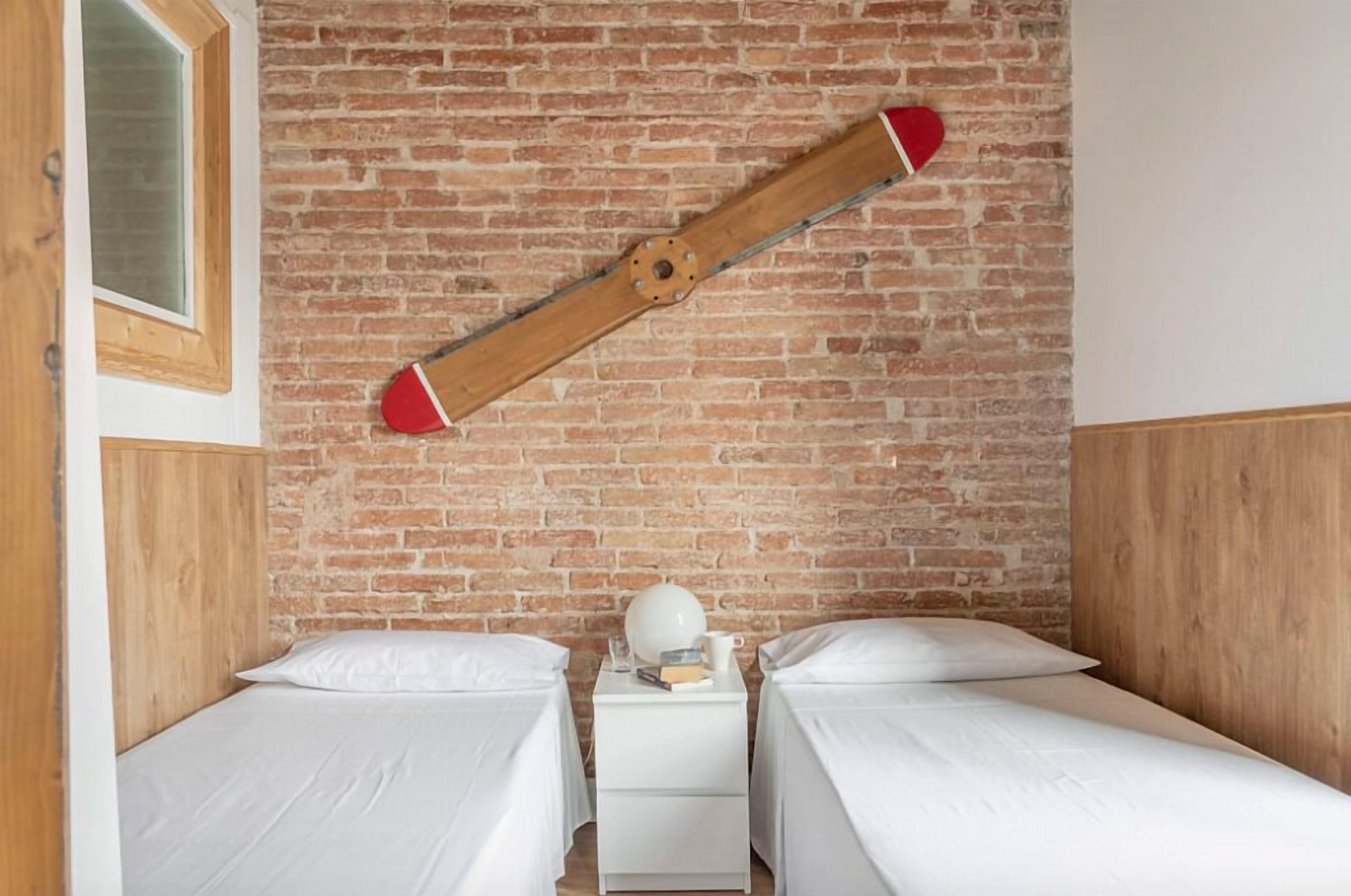 Een kamer met twee bedden, een houten hoofdbord, een bakstenen muur en een nachtkastje van hout met een witte lamp en boeken.