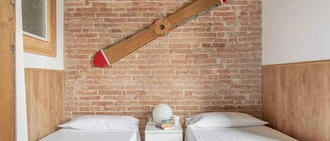 Een kamer met twee bedden, een houten hoofdbord, een bakstenen muur en een nachtkastje van hout met een witte lamp en boeken.