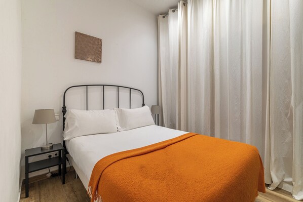 A neatly made bed with white bedding and an orange throw blanket, a black metal headboard, a bedside table with a lamp, and a white curtain.