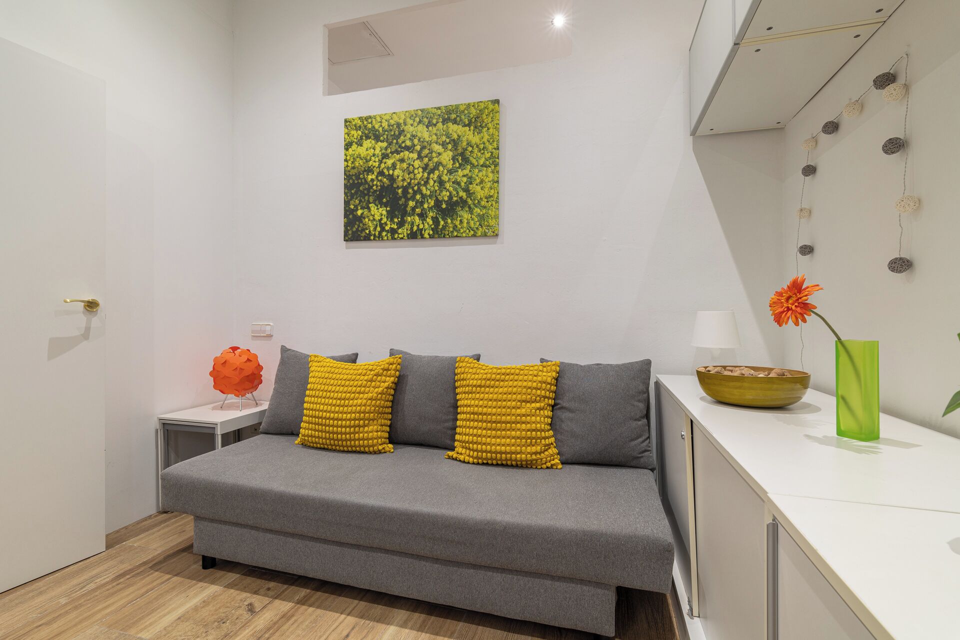 A modern living room with a grey sofa, two yellow pillows, a green vase with an orange flower, and a painting on the wall.