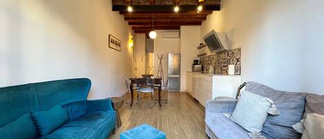 A compact living space with a blue sofa, a grey sofa, a dining table with chairs, and a kitchenette.