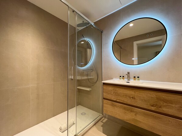 Un baño moderno con ducha envuelta en vidrio, tocador de madera y espejo redondo con iluminación ambiental.