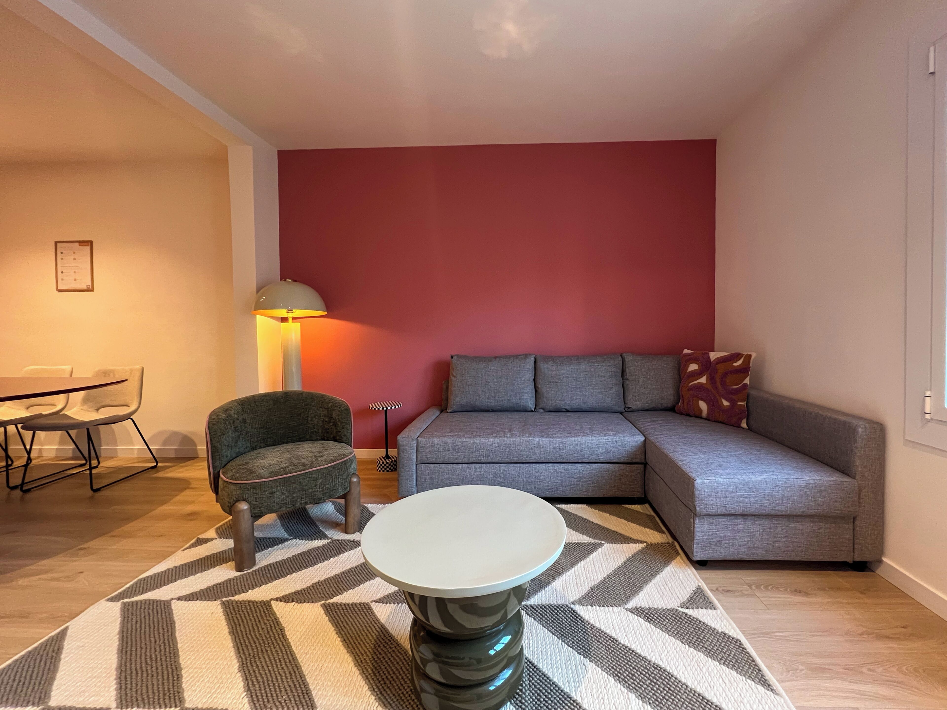 Una sala de estar moderna con un sofá modular, una mesa de centro redonda, una alfombra estampada, una lámpara y un comedor con sillas y una mesa.