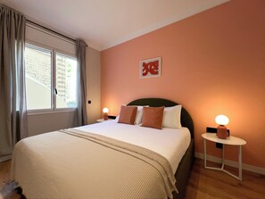 A hotel room with a bed, two bedside tables, a window with curtains, a wall with a red sign, and a small table with a lamp.