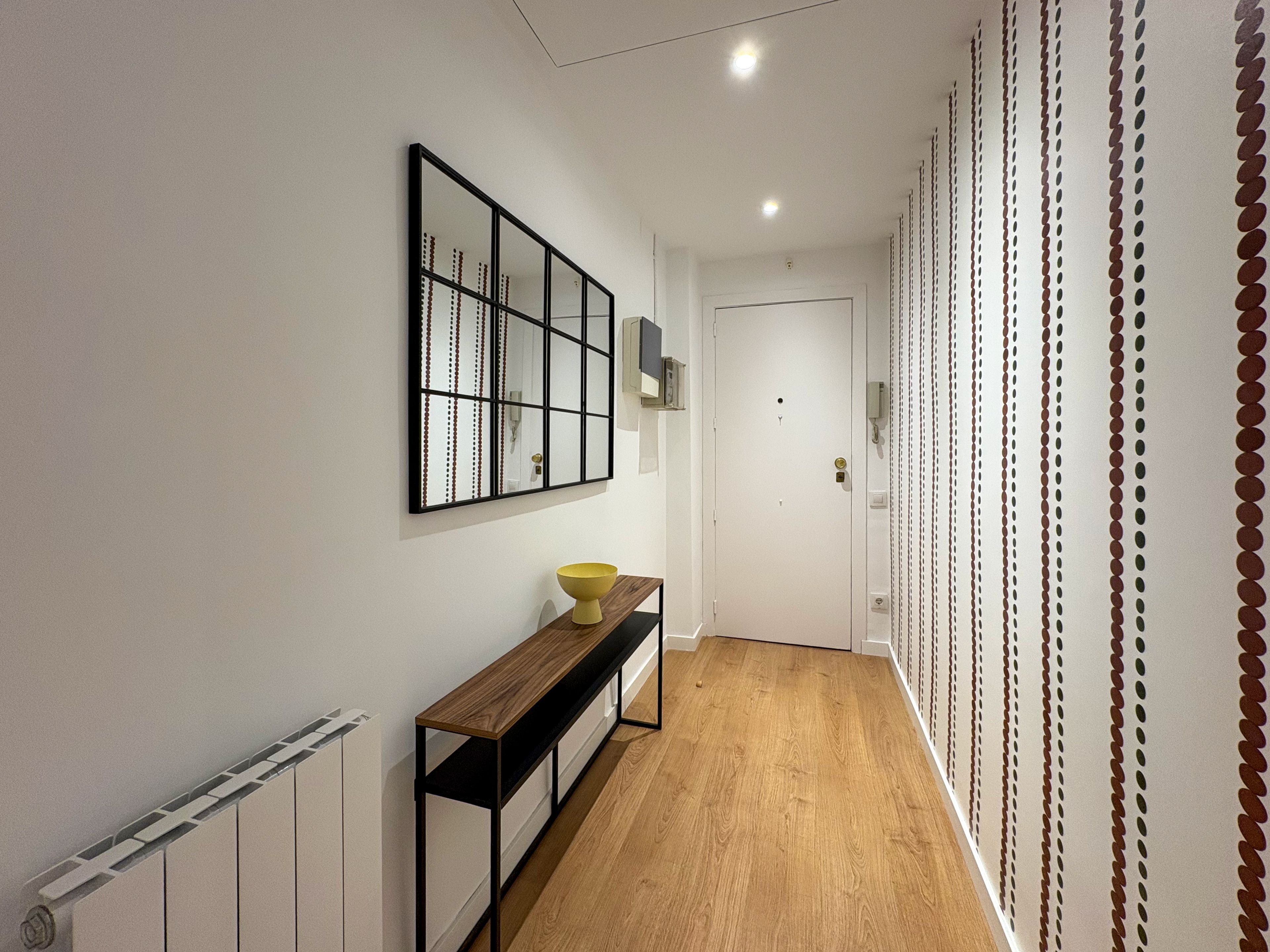 Un pasillo con suelo de madera, un banco, un espejo en la pared y una puerta.