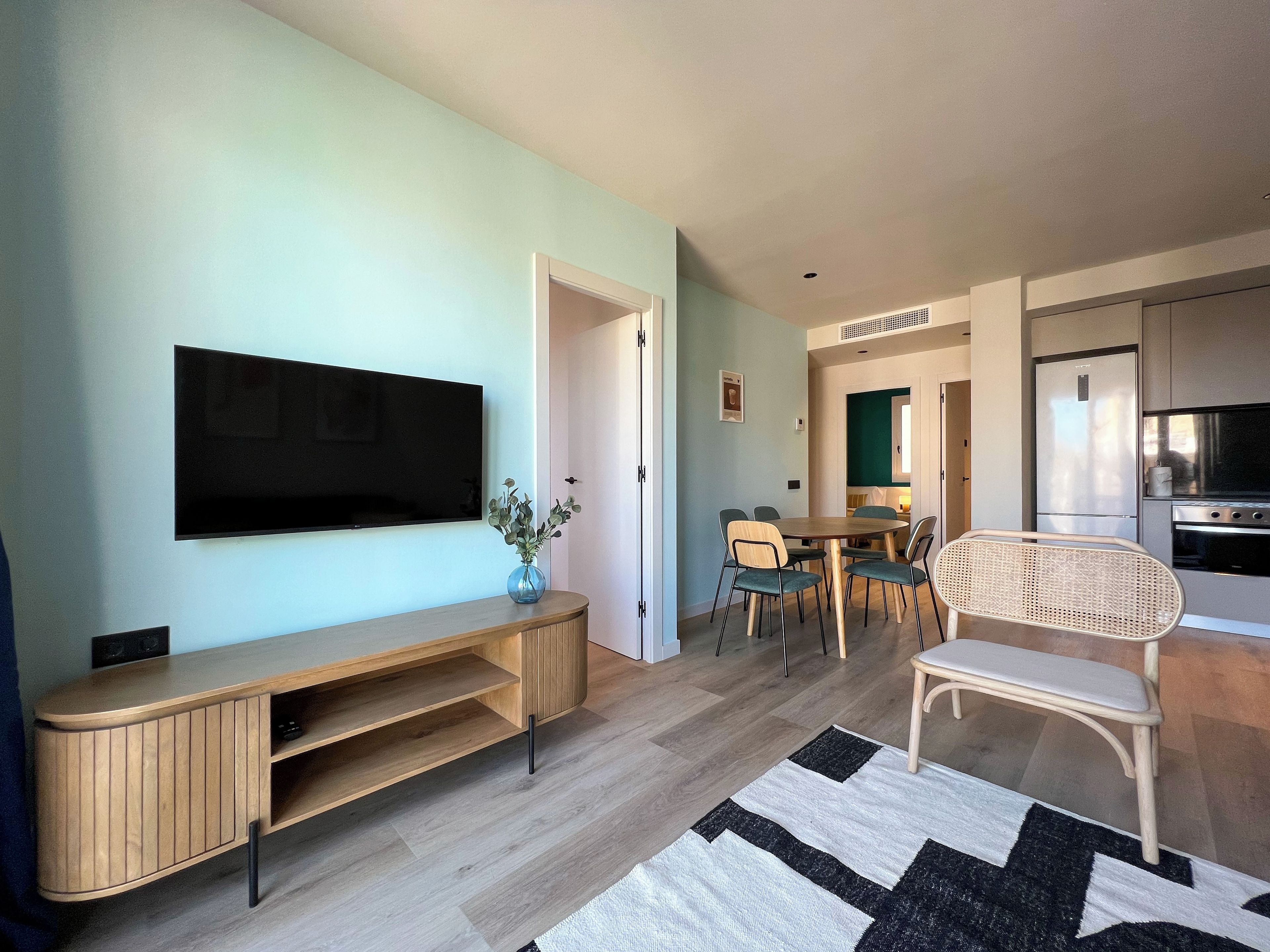 Una sala de estar moderna con televisor de pantalla plana, mueble de madera para entretenimiento, mesa de comedor con sillas y una zona de cocina con refrigerador y horno.
