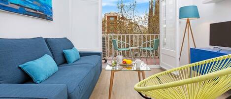 Un salon moderne avec un canapé bleu, un fauteuil jaune, une petite table sur laquelle se trouvent des fruits, et un balcon équipé de chaises et d’un téléviseur.