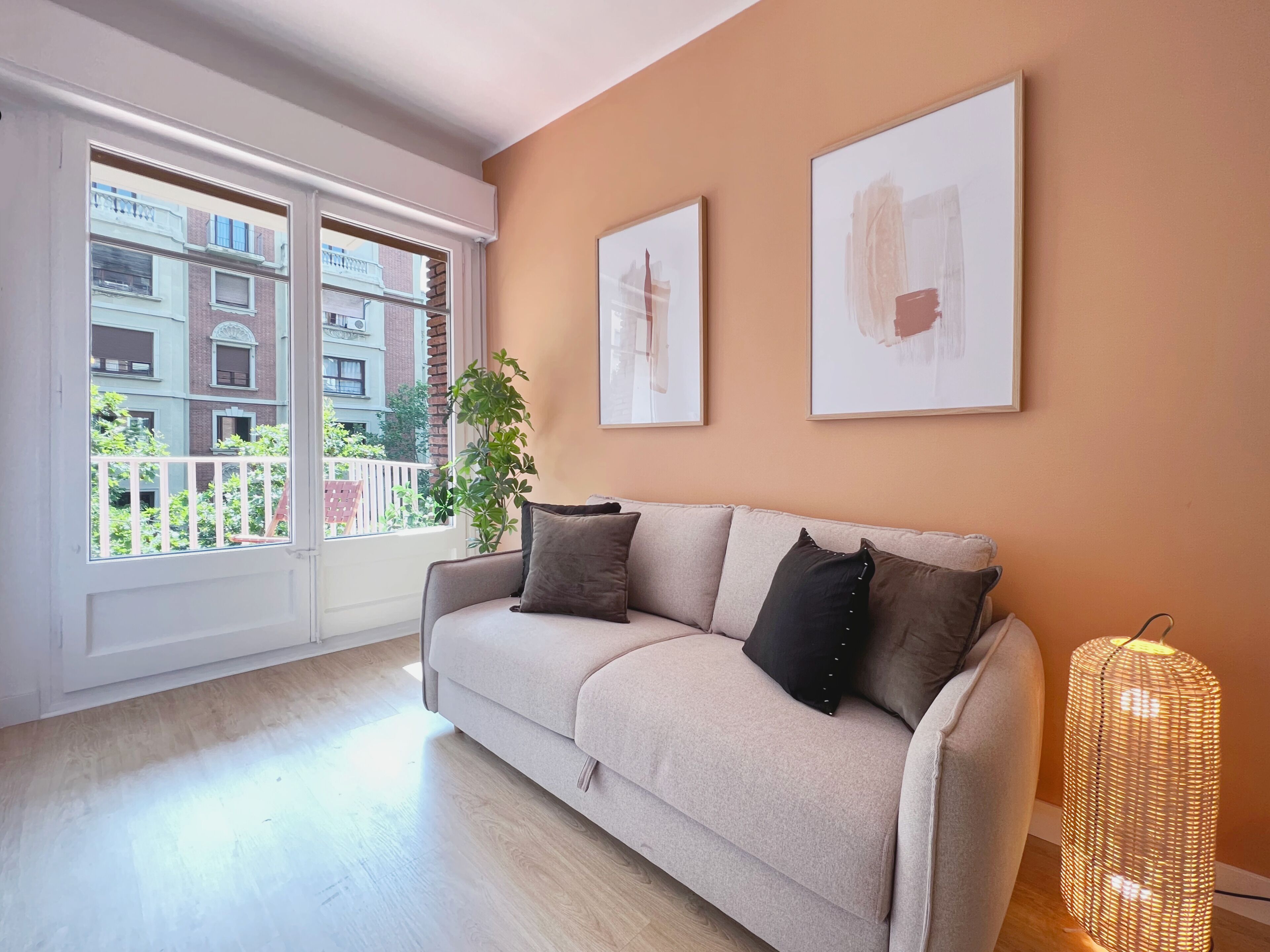 A living room with a beige sofa, two framed artworks, a wicker floor lamp, and a view of buildings through a large window.