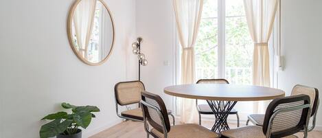 A dining area with a round table and four chairs, a potted plant, a mirror, and a window with curtains.