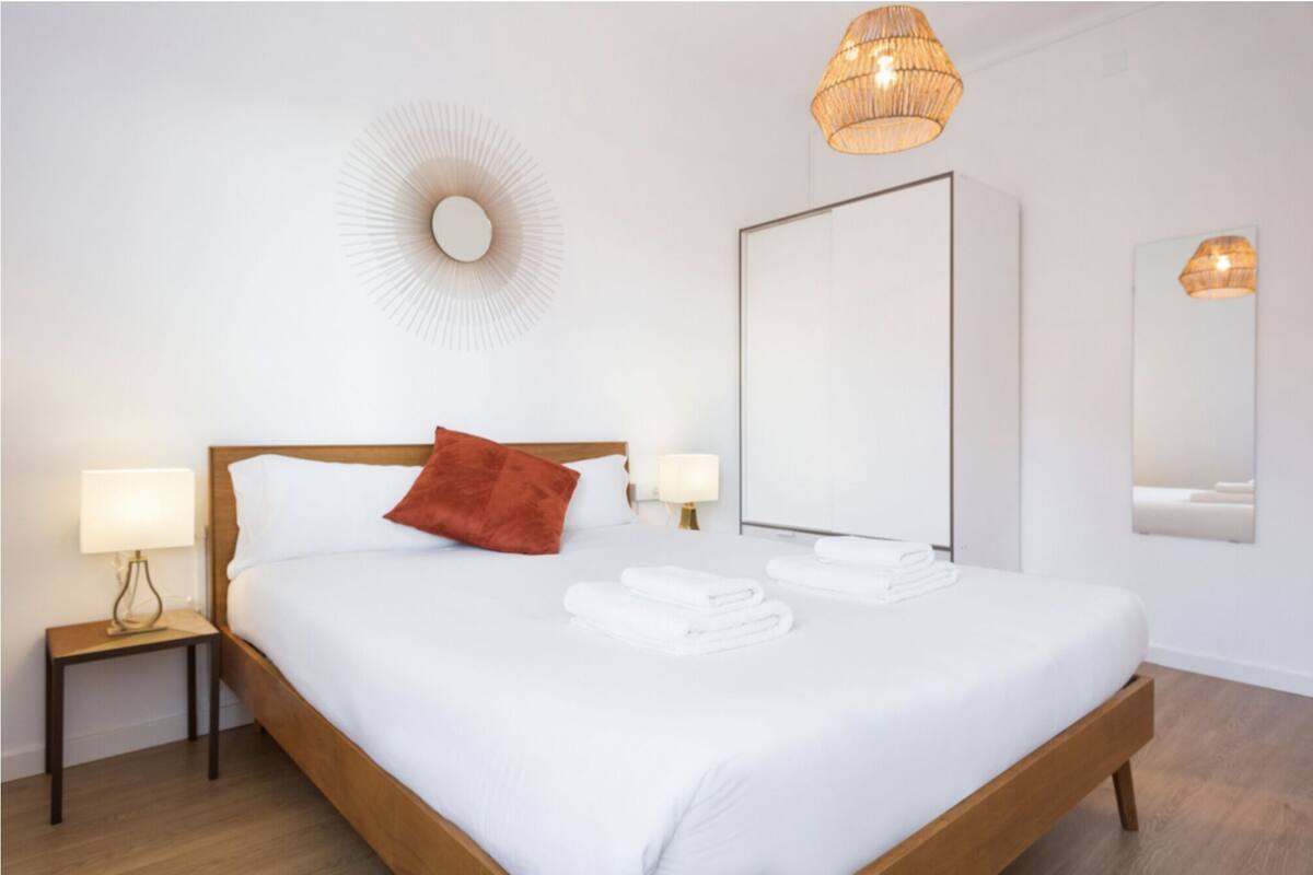 Un lit bien fait, avec du linge de lit blanc, une tête de lit en bois, un coussin rouge et un élément décoratif rond fixé au mur.