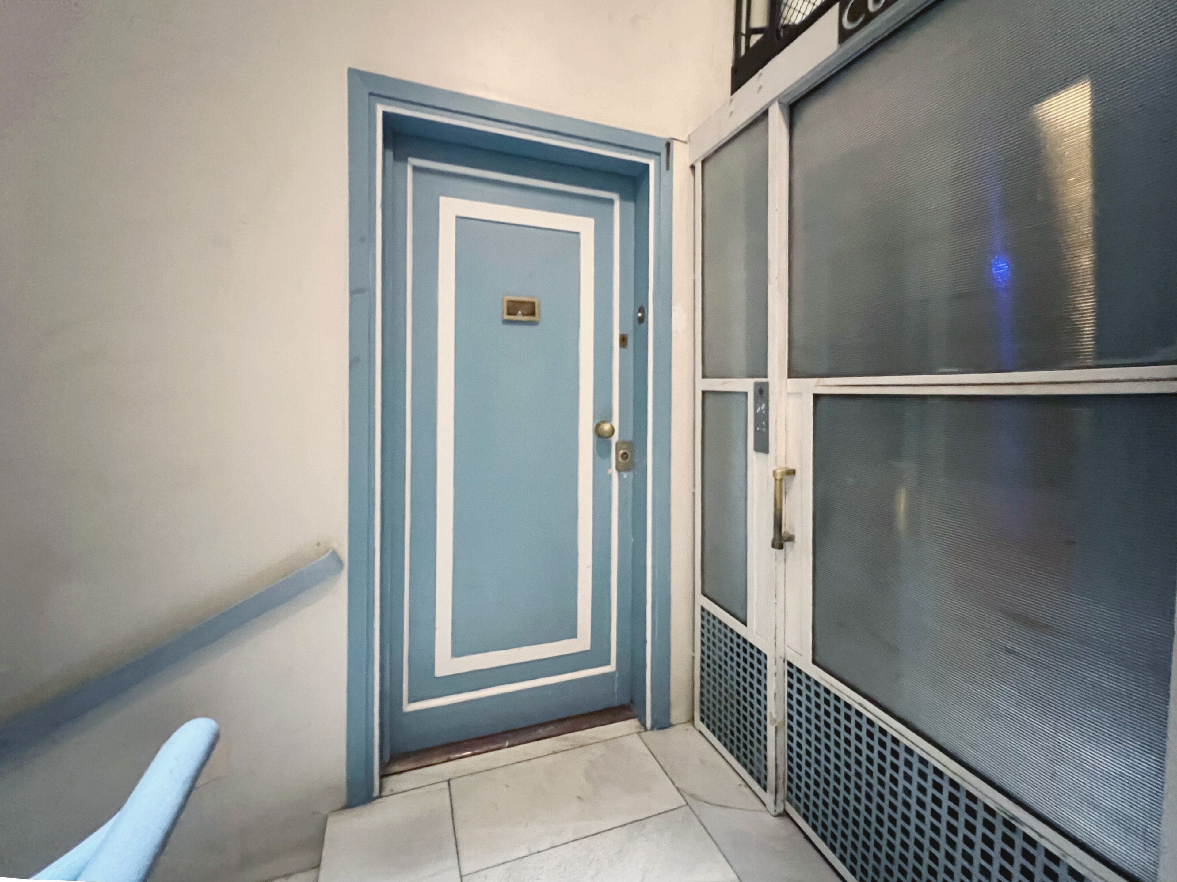 Una puerta azul con picaporte de metal y una placa, una reja metálica con mosquitero y un suelo de baldosas.