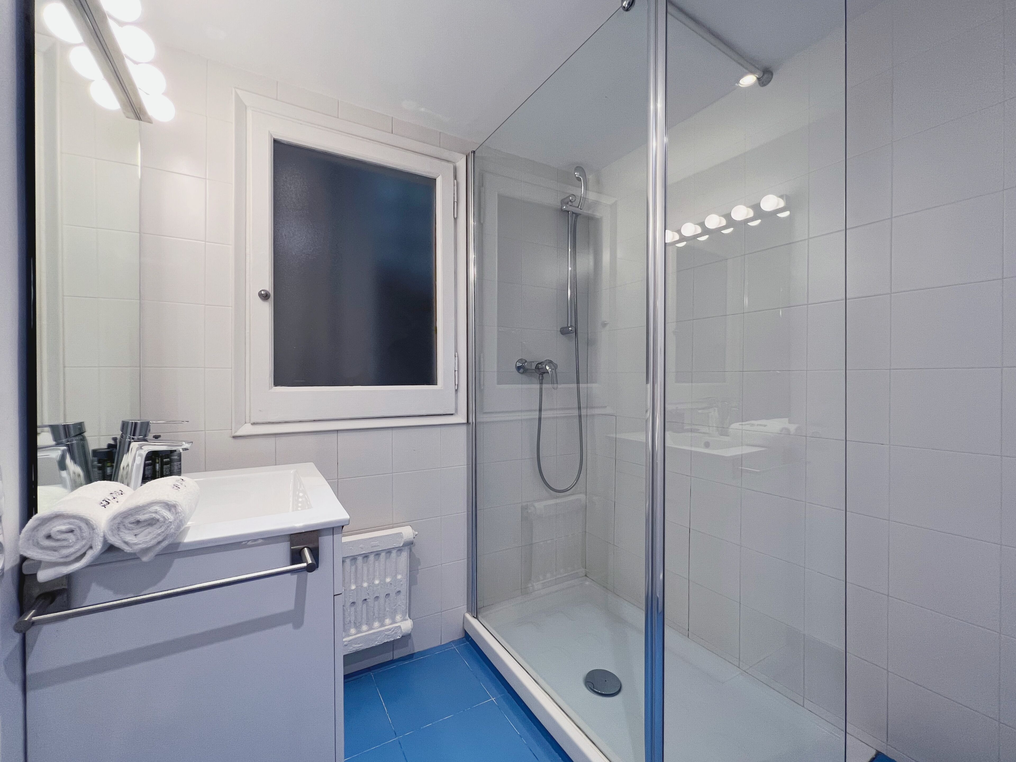A modern bathroom with a glass shower enclosure, a white sink with a towel rack, and a window above the mirror.