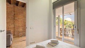 A bedroom with a large bed, a brick wall, and a sliding glass door leading to an outdoor patio with chairs. - Stay U-nique Apartments Nació (Barcelona)