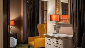 Camera d'albergo con un letto, una poltrona gialla, una cassettiera bianca, uno specchio e una lampada rossa.