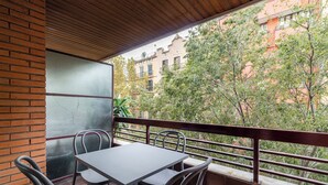 Uma varanda com mesa e cadeiras, vista para prédios e árvores, e um muro de tijolos.