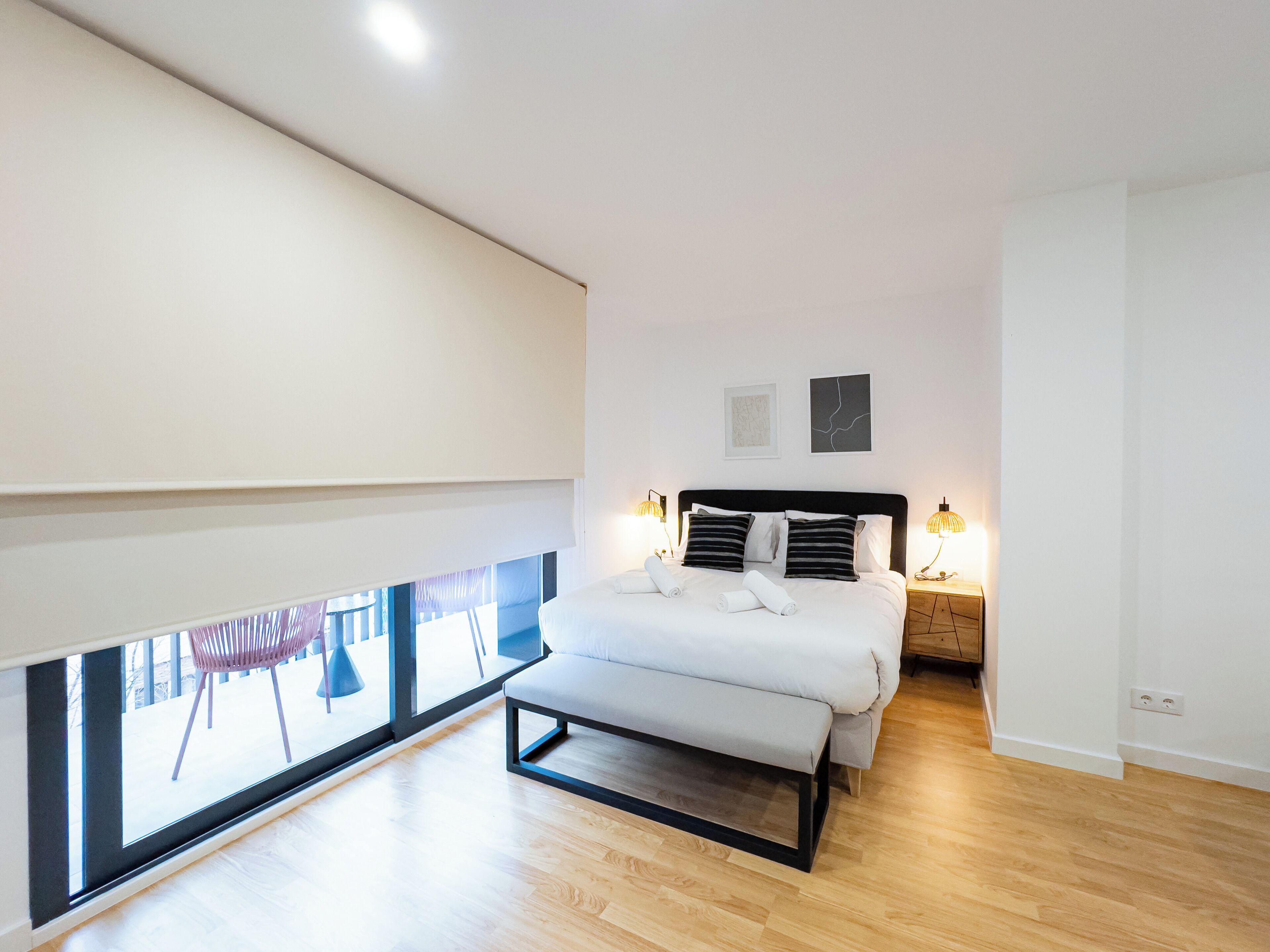 Un dormitorio moderno con un ventanal grande, una cama con reposabajera, lámparas de noche y vistas al exterior.