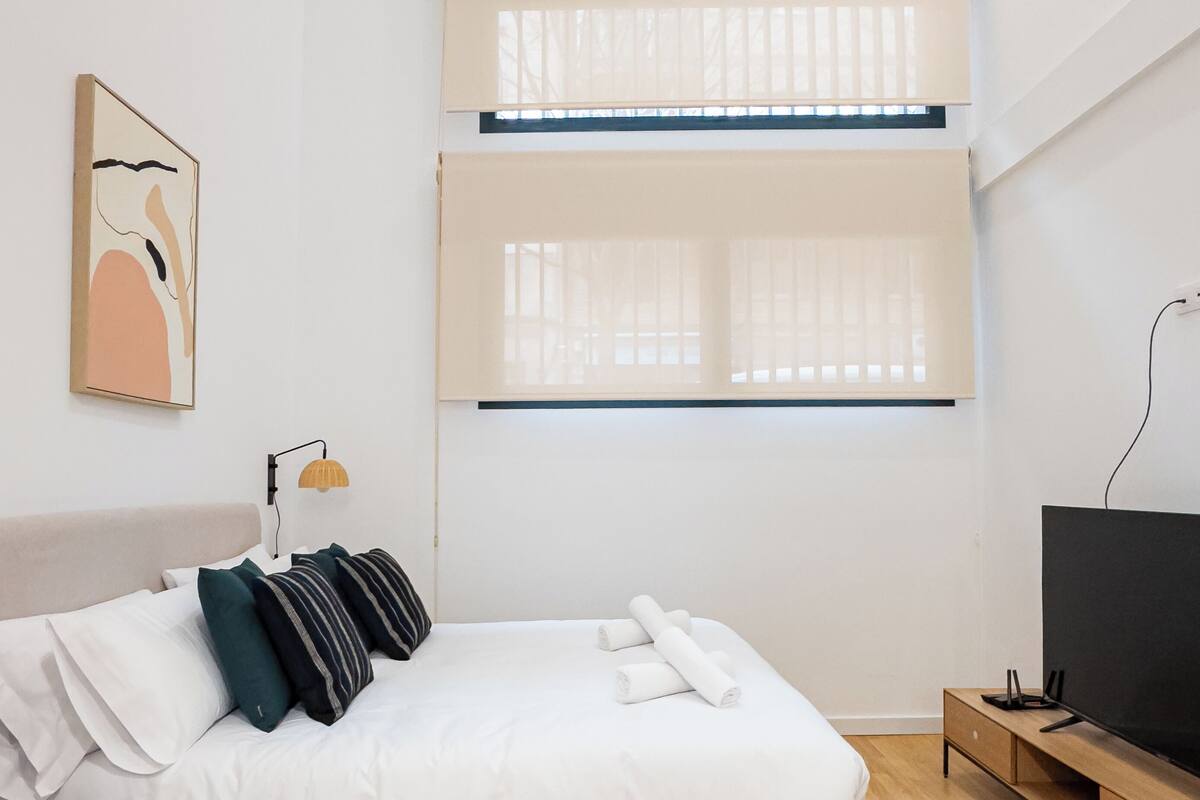 A modern bedroom with a bed, a TV, and a window with blinds.