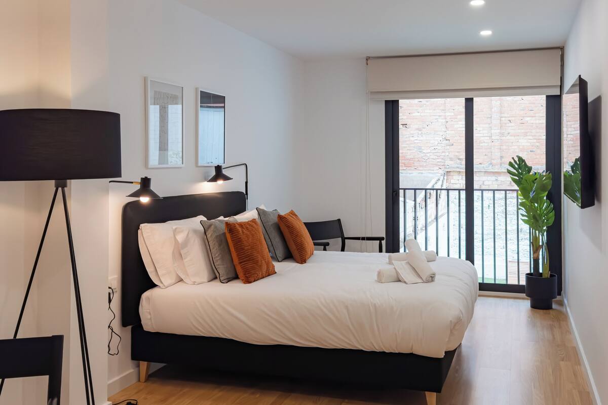 A modern bedroom with a black bed, white bedding, and a large window with a view of a brick building.