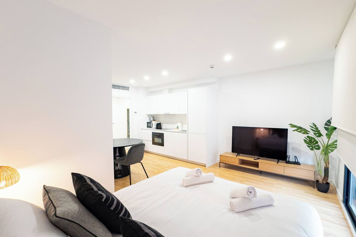 A modern hotel room with a large bed, a TV, a dining area, and a plant.