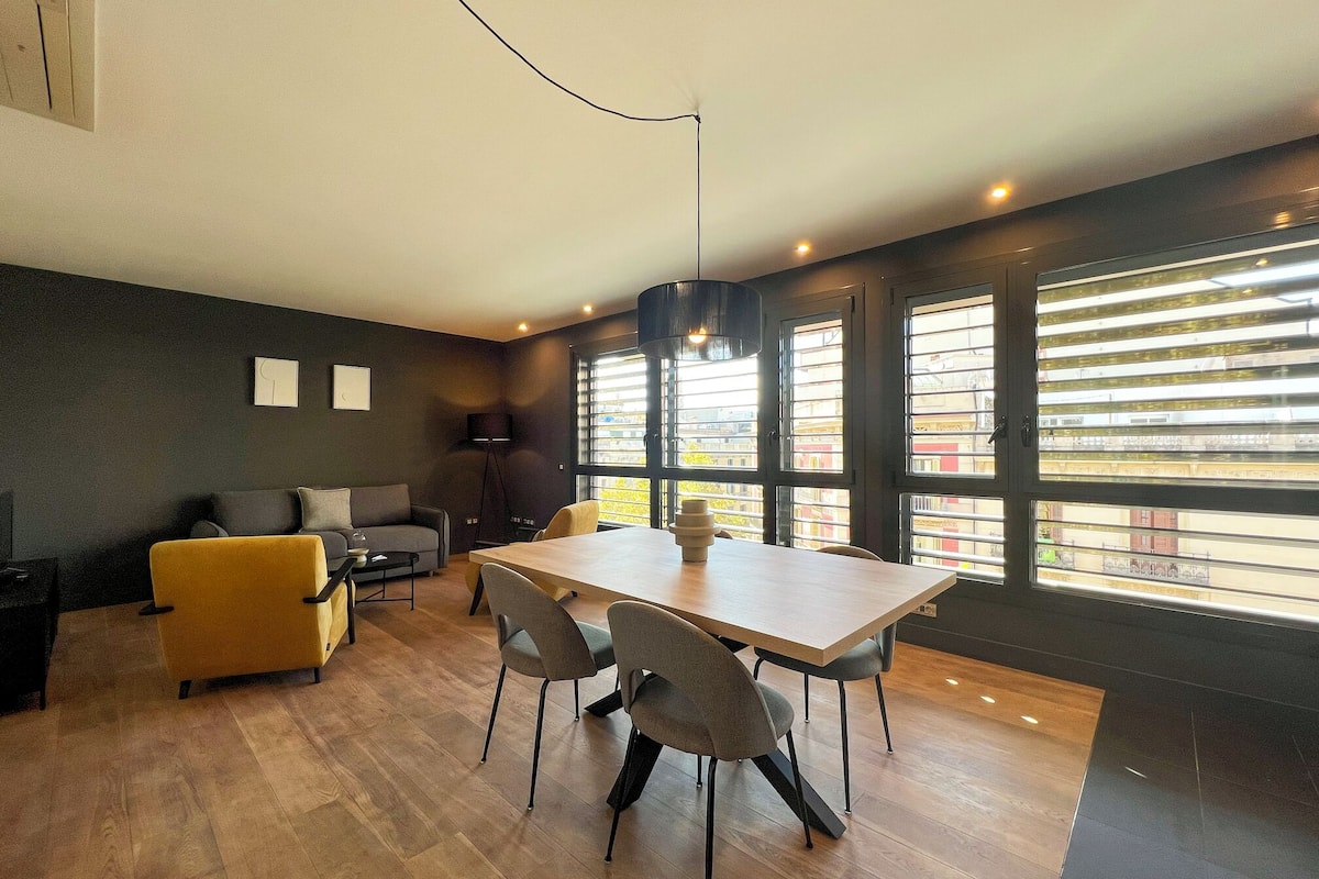 A modern living room with a dining area, featuring a large window with blinds, a sofa, a yellow armchair, and a dining table with chairs.