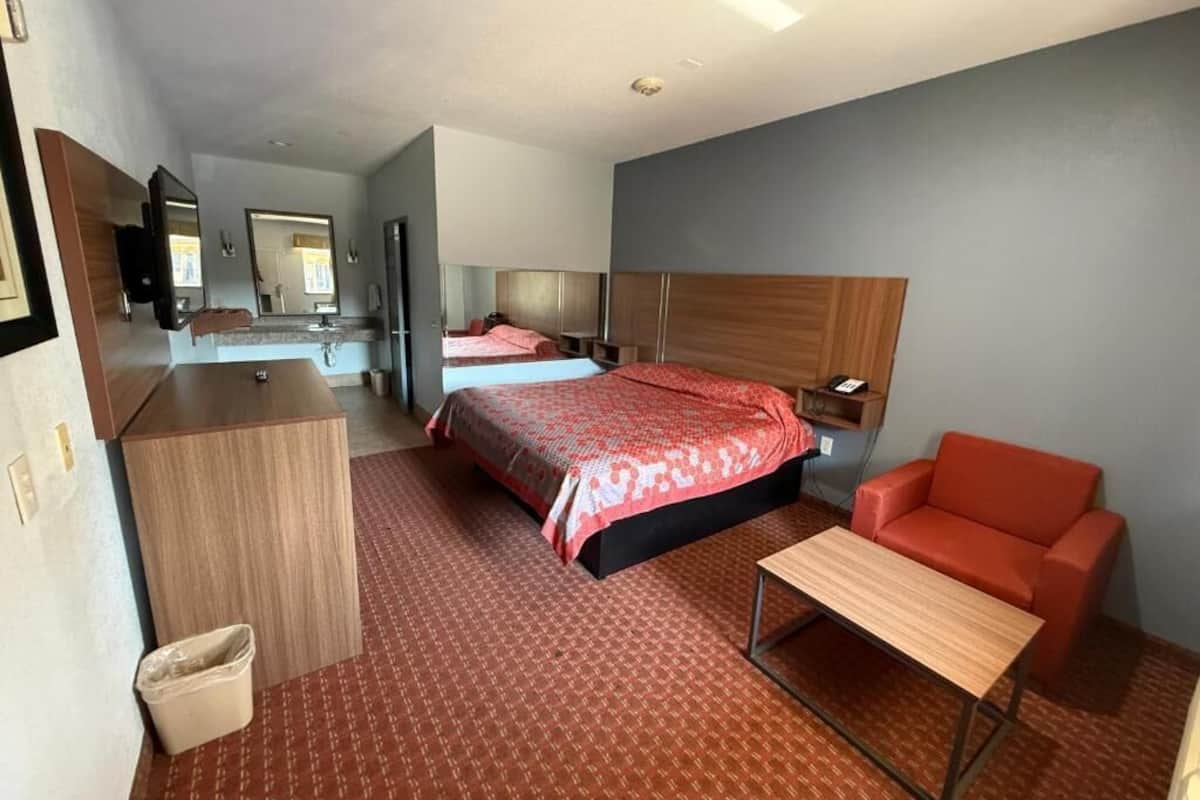 A hotel room with a large bed, a desk, a chair, a small table, a trash bin, and a mirror.