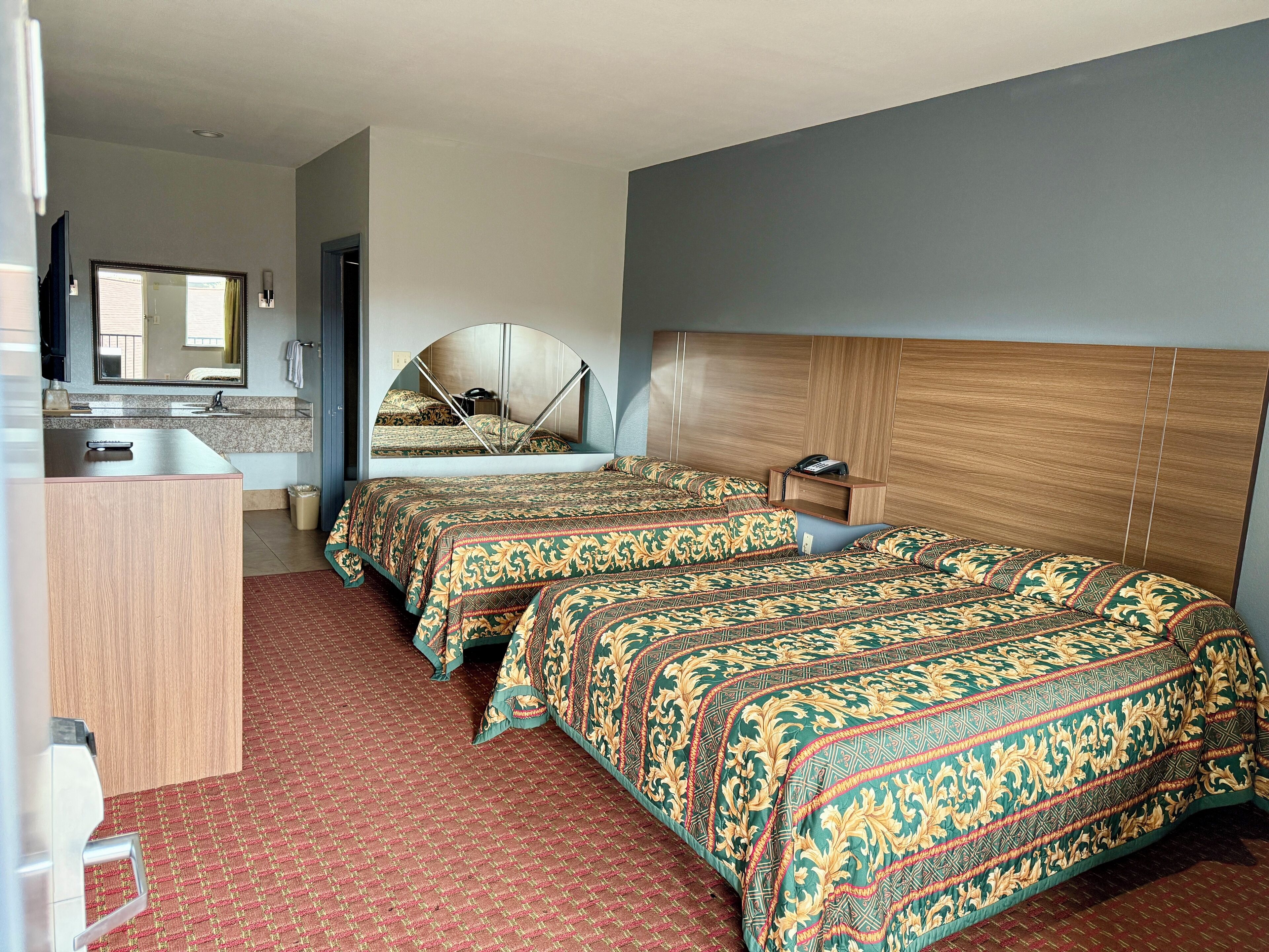 A hotel room with two beds, a wooden headboard, a mirror, a small bedside table, and a desk with a chair.