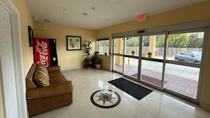 A lobby with a Coca-Cola sign, a brown leather couch, potted plants, and a large glass door leading outside. - Budget Host Inn Stafford - Houston (Houston)
