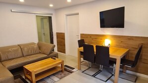 Ein Wohnzimmer mit einer Couch, einem Holztisch, einem Esstisch mit Stühlen, einem an der Wand montierten Fernseher und einer Tür zu einem anderen Raum.
