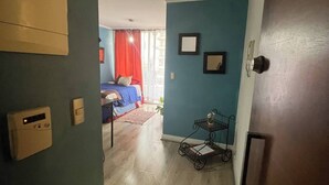 A room with a bed, a small table, and a chair. There is a window with red curtains and a view of the outside.