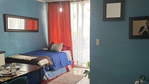 A bedroom with a bed, a desk, and a chair. There is a window with curtains, a mirror, and a framed picture on the wall.