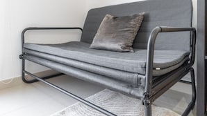 Un canapé gris avec un coussin et un oreiller, une structure en métal et un tapis tissé sur le sol.
