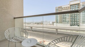 Ein Balkon mit zwei weißen Stühlen und einem kleinen Tisch, mit Blick auf eine Stadtlandschaft aus modernen Gebäuden und einer Autobahn.
