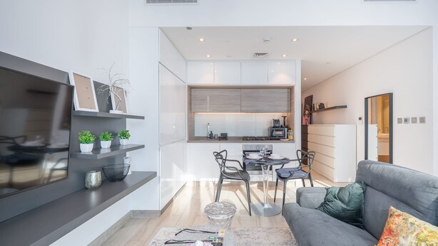 A modern living room with a grey sofa, a coffee table, and a TV on a wall. There is a kitchen area in the background with a dining table and chairs. - Dubai Marina Studio for 2 With Sea Views (Dubai)