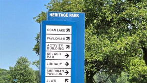 A directional sign at Heritage Park with arrows pointing to various facilities including Coan Lake, Pavilion A-B, Activity Building, Splash Pad, Library, Sheridan Pavilion, and JLWHS.