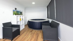 A room with a hot tub, two wicker chairs, a white shelf unit, and a wall-mounted TV.