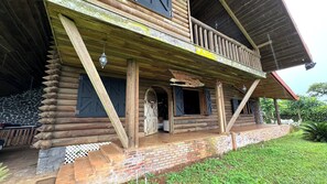 Een rustiek houten blokhut met een balkon, stenen treden en een fundering van baksteen.