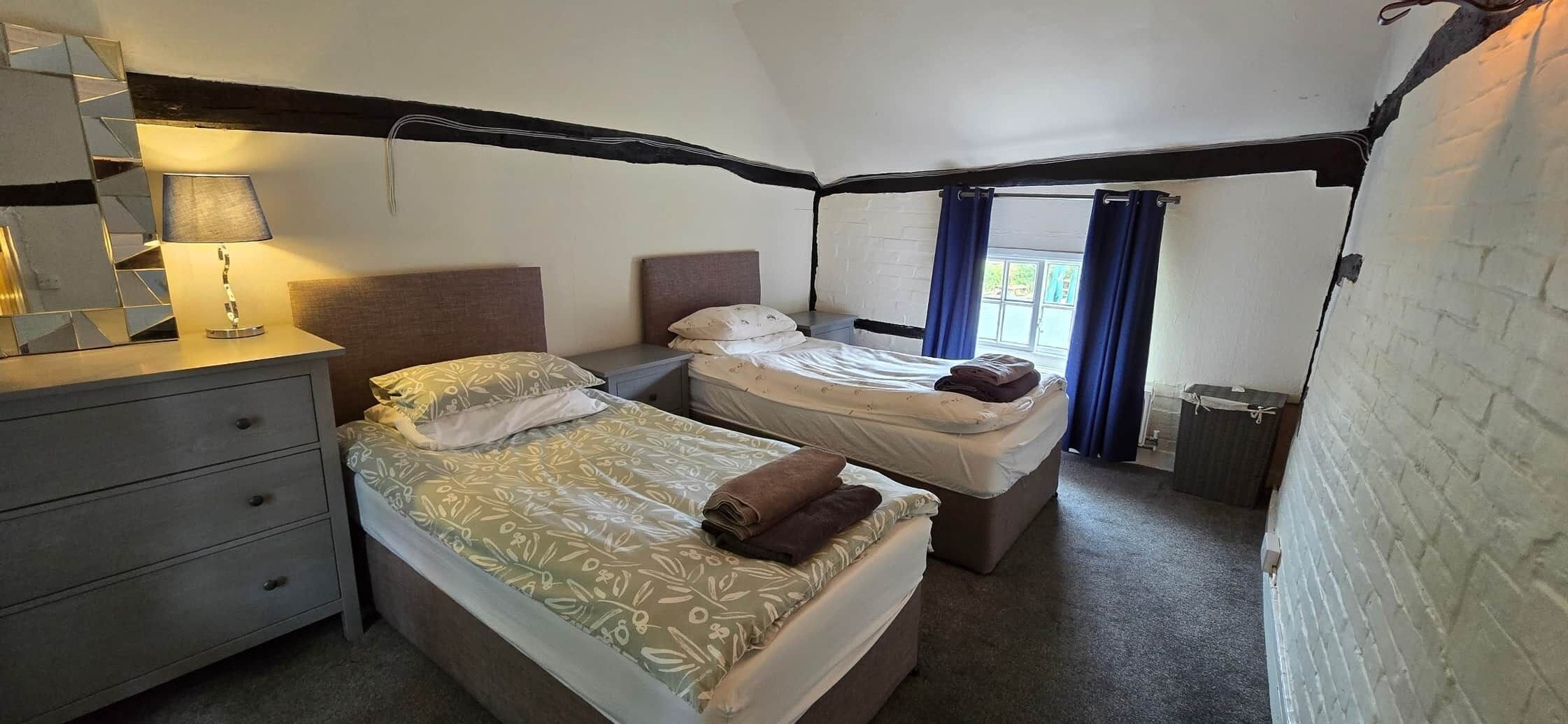 Ein Schlafzimmer mit zwei Betten, einem Nachttisch mit Lampe und einem Fenster mit Vorhängen.