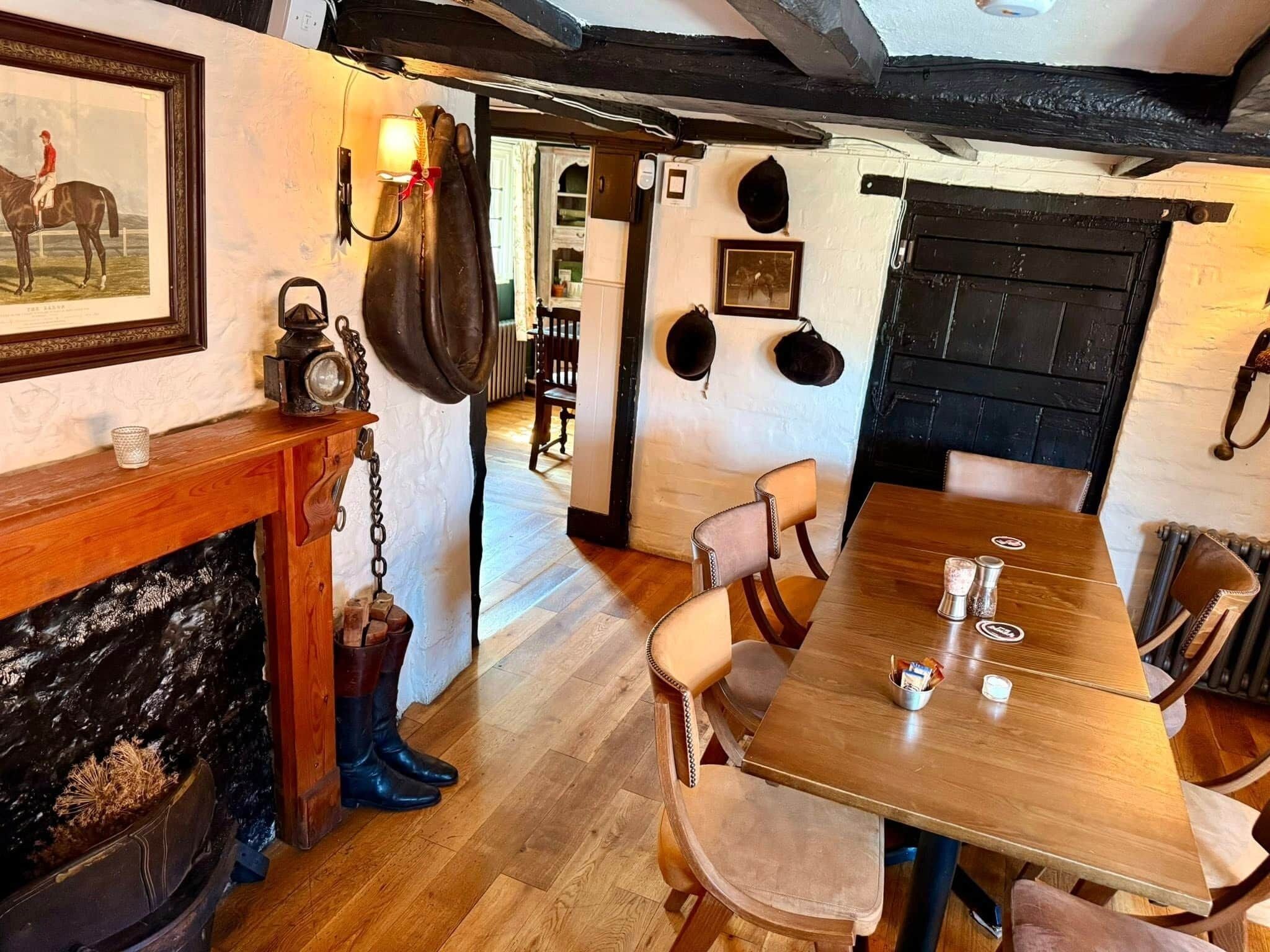 A wooden dining table with chairs, a fireplace with a mantle, and a wall with mounted hats and a framed picture.