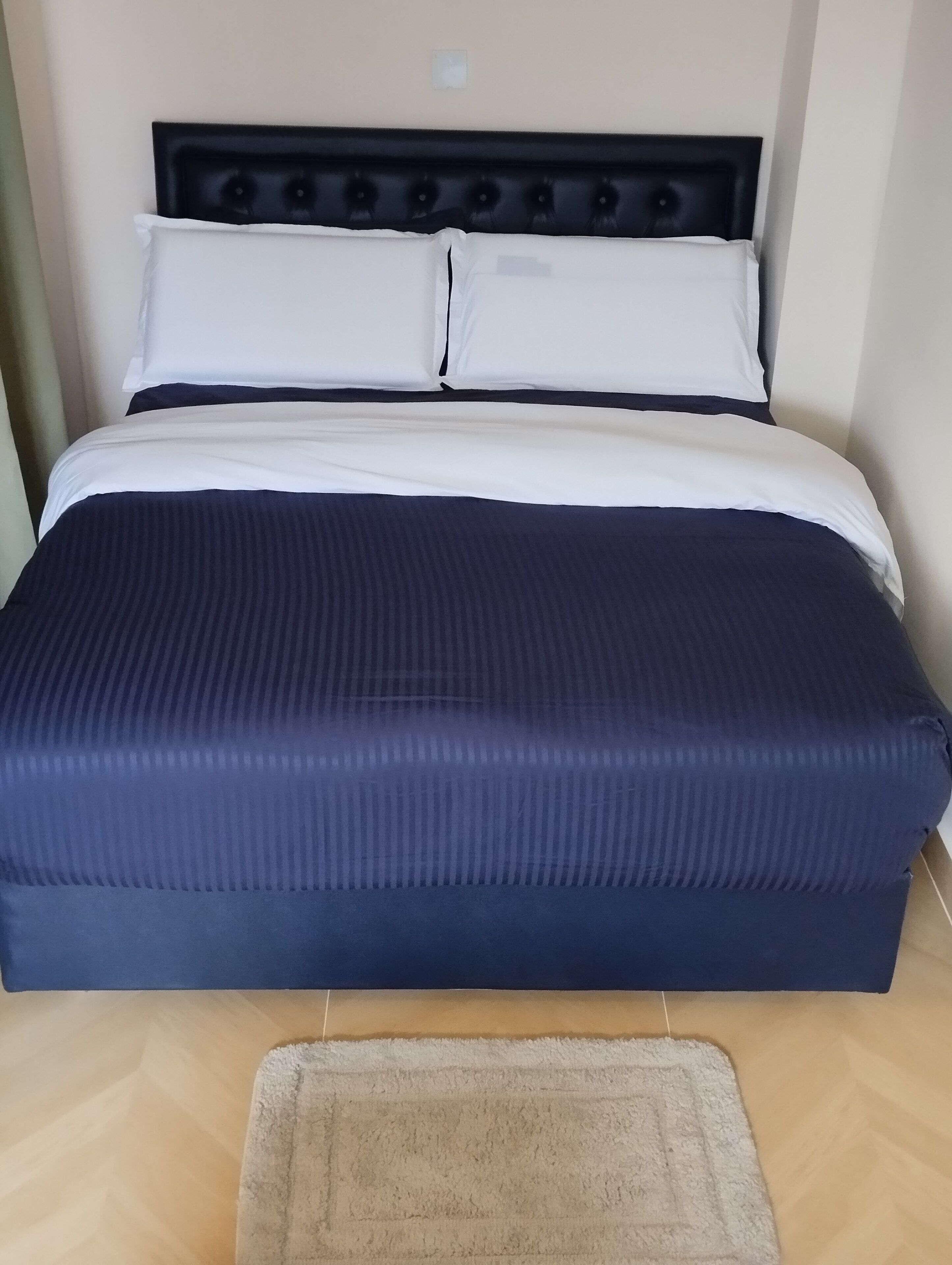Un lit avec un matelas bleu foncé et des oreillers blancs, une tête de lit à motifs capitonnés et un tapis de couleur claire sur le sol.