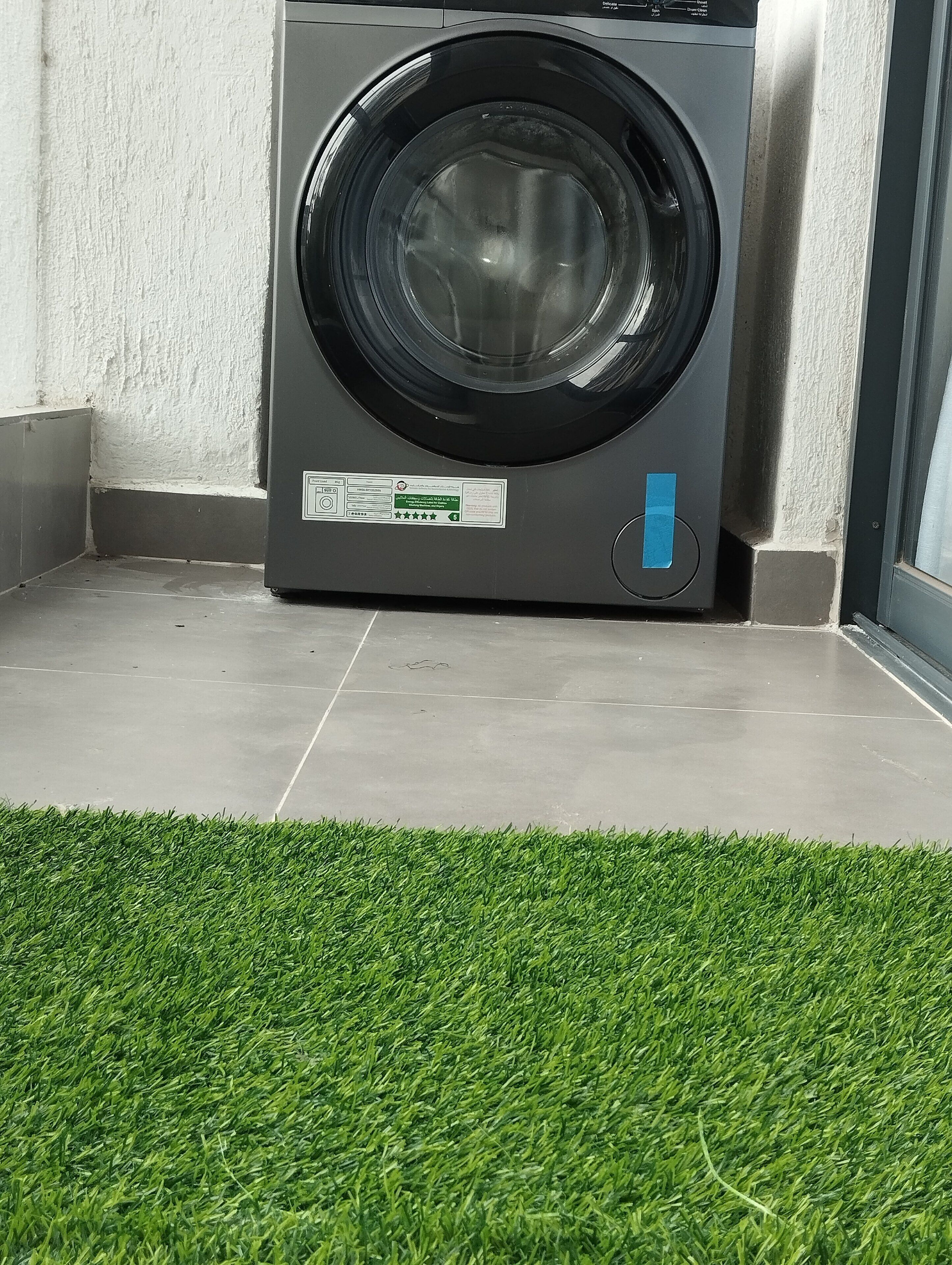 A front-loading washing machine in a corner with a small patch of artificial grass in front.