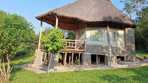 Une cabane à toit de chaume, dotée d’un porche en bois et d’une base en béton, entourée de verdure.