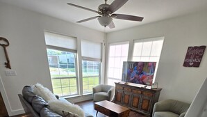 Una sala de estar con un sofá gris, una mesa de centro de madera y un televisor sobre un mueble de madera. Hay dos ventanas con persianas, un ventilador de techo y un elemento decorativo en la pared.