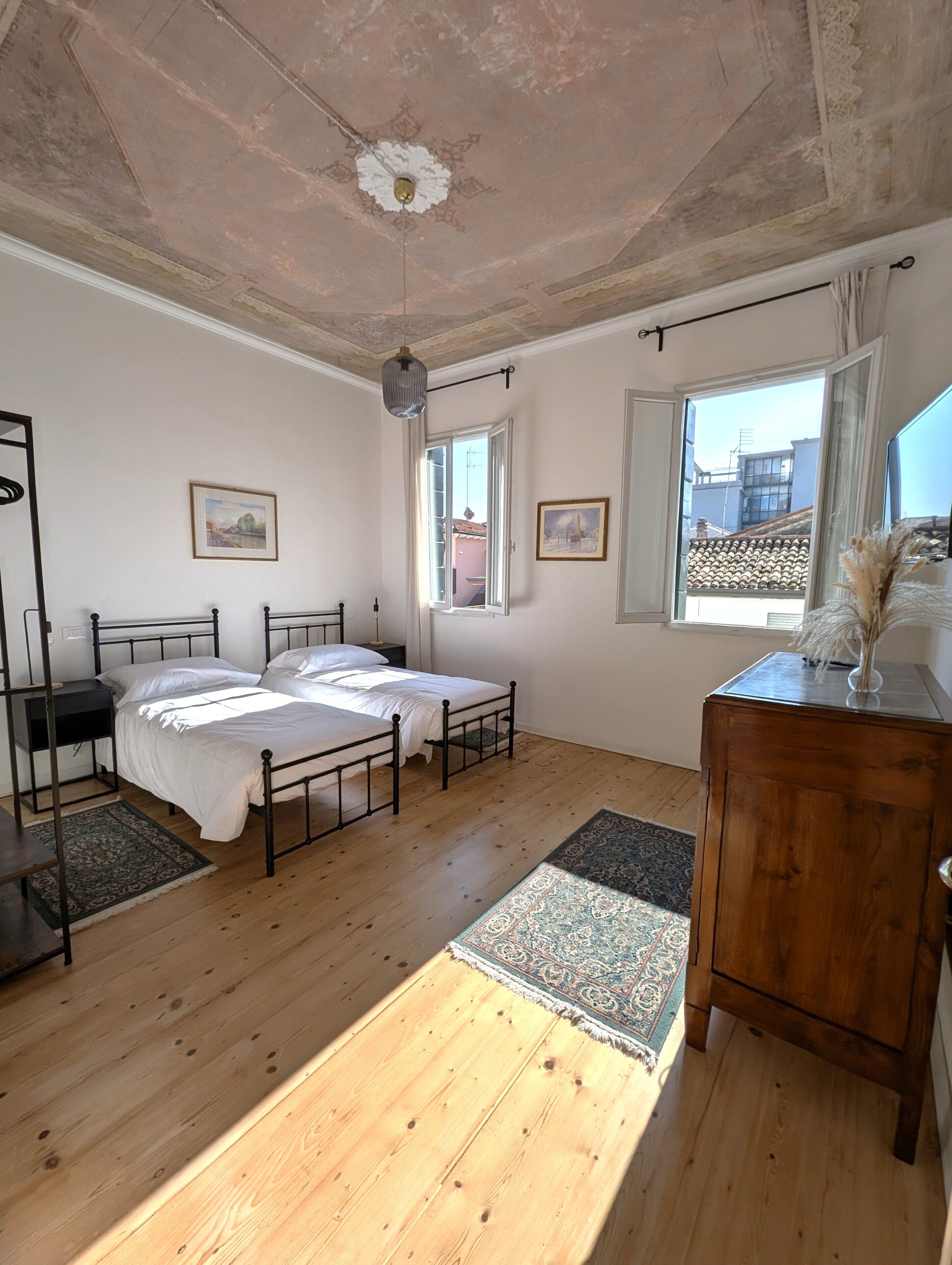 Ein Schlafzimmer mit zwei Betten, einem Holzschrank, einem Nachttisch, einem Deckenventilator und einem Fenster mit Blick auf Gebäude.