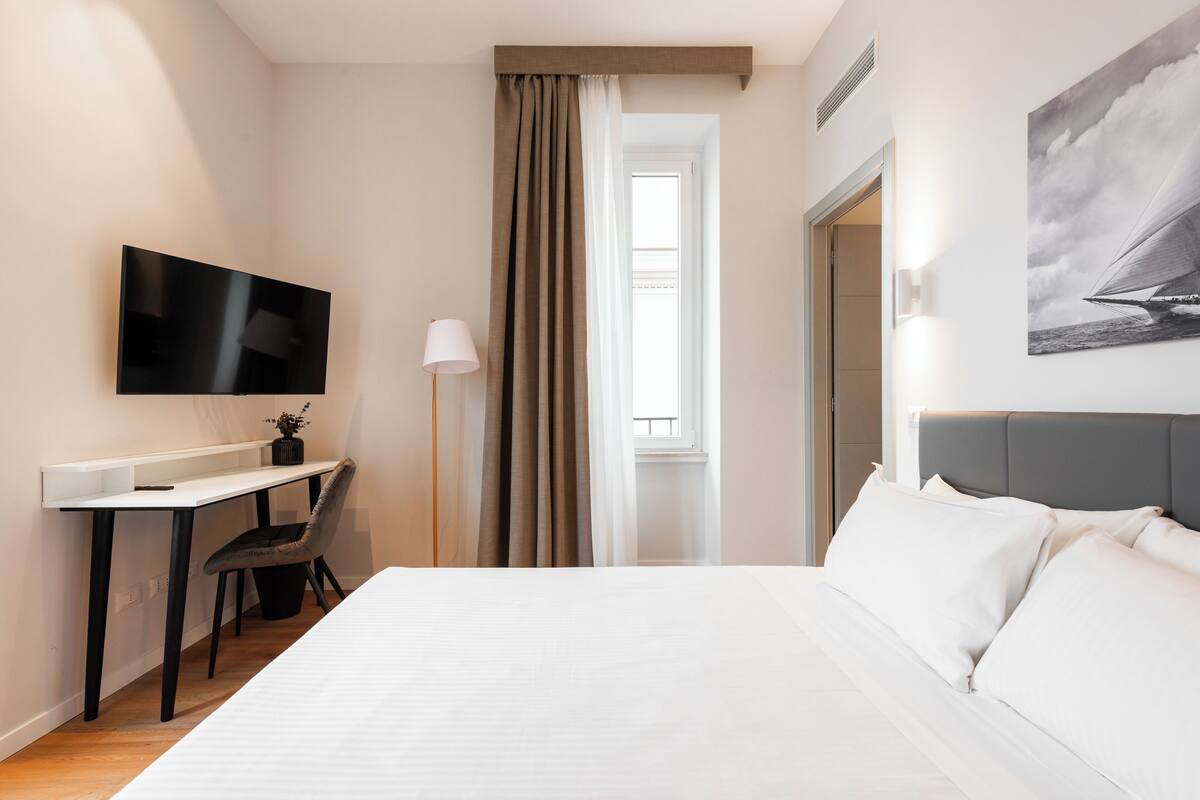 Habitación de hotel con cama, escritorio con silla, televisión, ventana con cortinas y un cuadro en la pared.