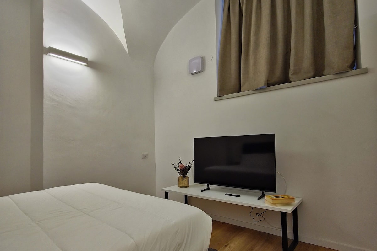 Un dormitorio con cama, televisor sobre un mueble, ventana con cortinas y una lámpara fijada a la pared.